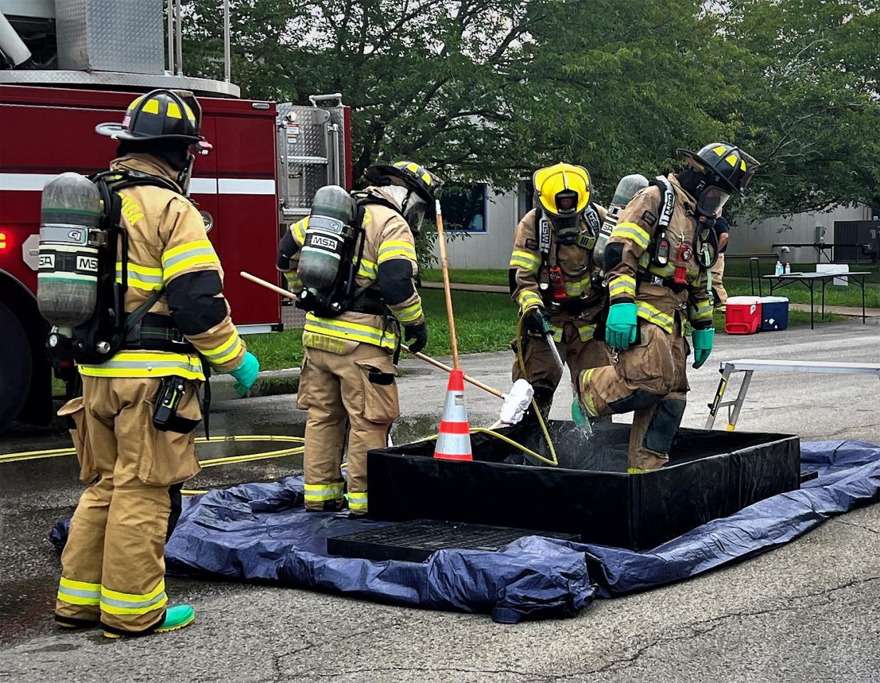 Four firefighters in full gear and breathing apparatus participate in a decontamination drill near a fire truck. One firefighter steps into a cleaning bin while others assist.