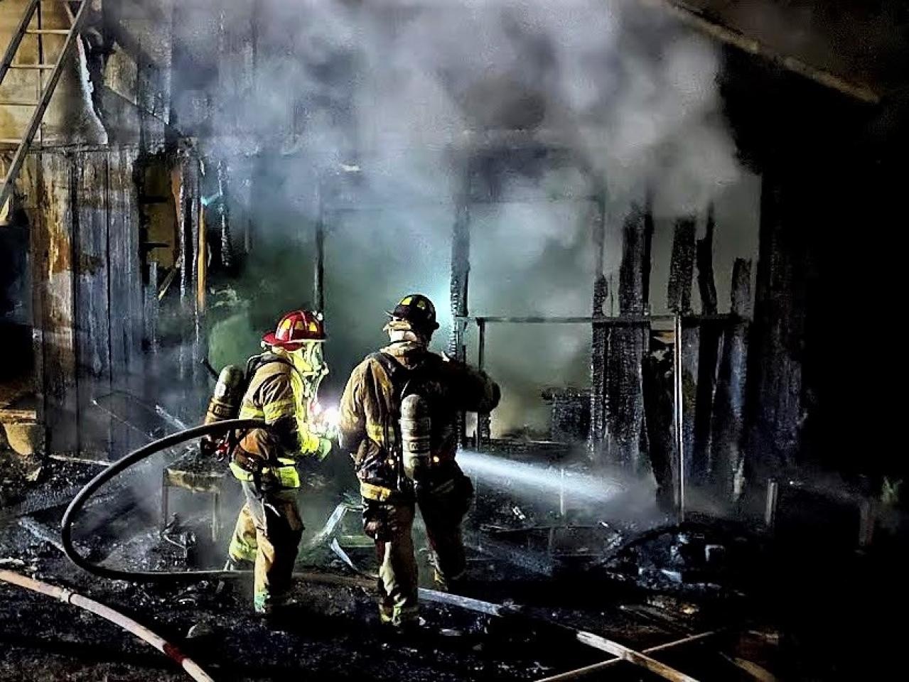 Two firefighters in full gear extinguish a nighttime blaze at a charred, smoking structure, illuminated by their helmet lights.