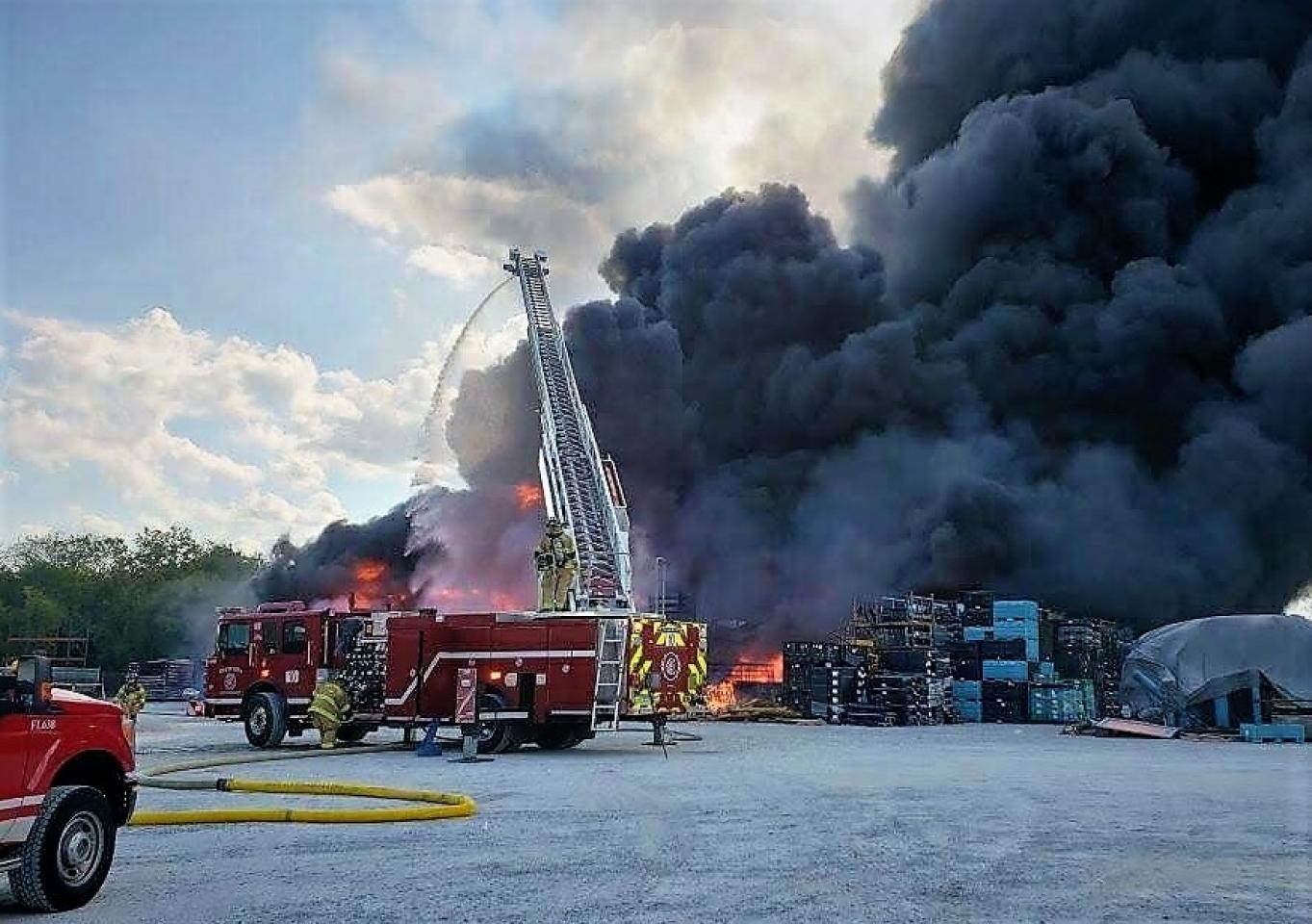 Firefighters combat a large fire at an industrial site with thick black smoke, using a ladder truck and hoses.