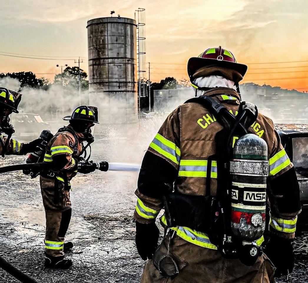 Firefighters in tan gear extinguishing a fire in an outdoor setting, with a large tower and smoke in the background at sunset