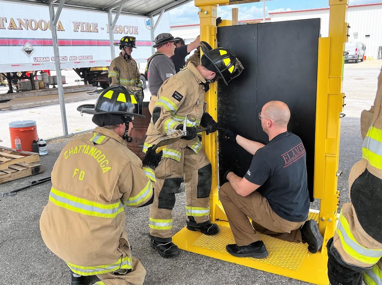 Firefighters in tan gear using tools to breach a door, supervised by an instructor in a black shirt, with a yellow frame in an outdoor training area.