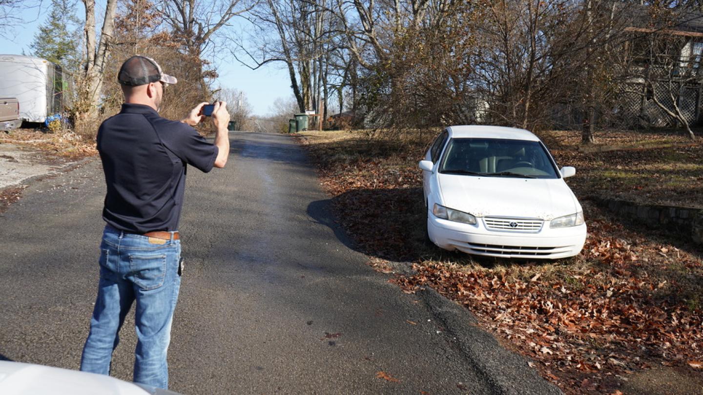 Man in a black shirt and jeans taking a photo of a white car parked on the side of a leaf-strewn road, with bare trees in the background.