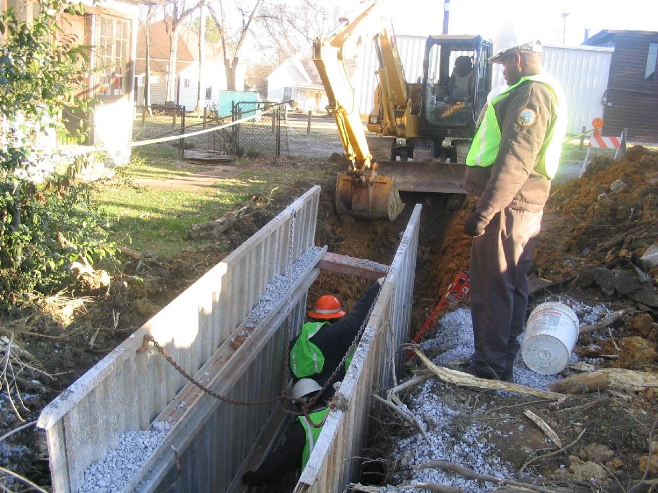 Two construction workers in green safety vests, one inside a metal trench box and another standing outside, with a yellow excavator in the background.