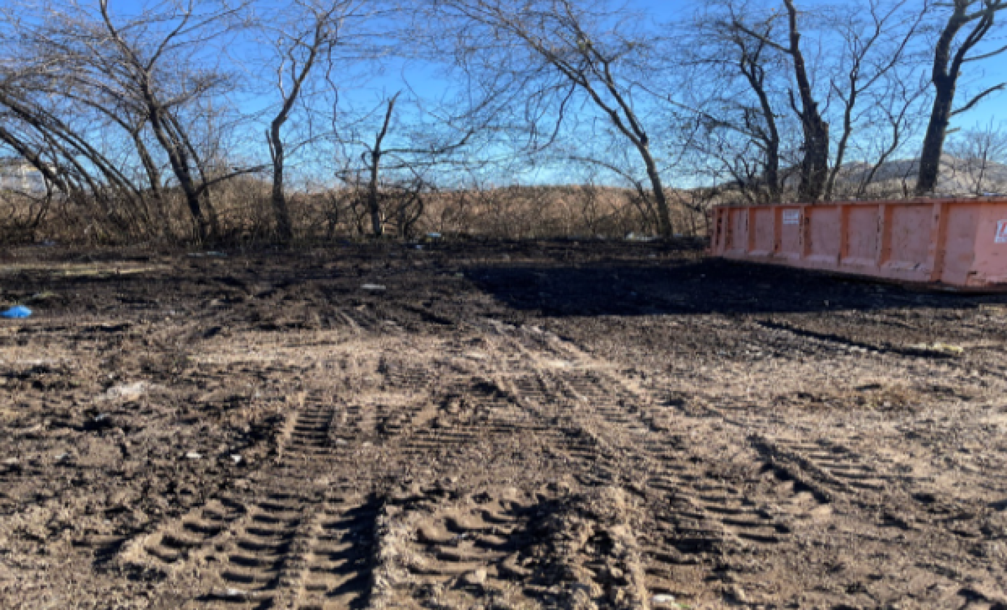 Cleared area with visible tire tracks on muddy ground, surrounded by leafless trees and a large orange dumpster