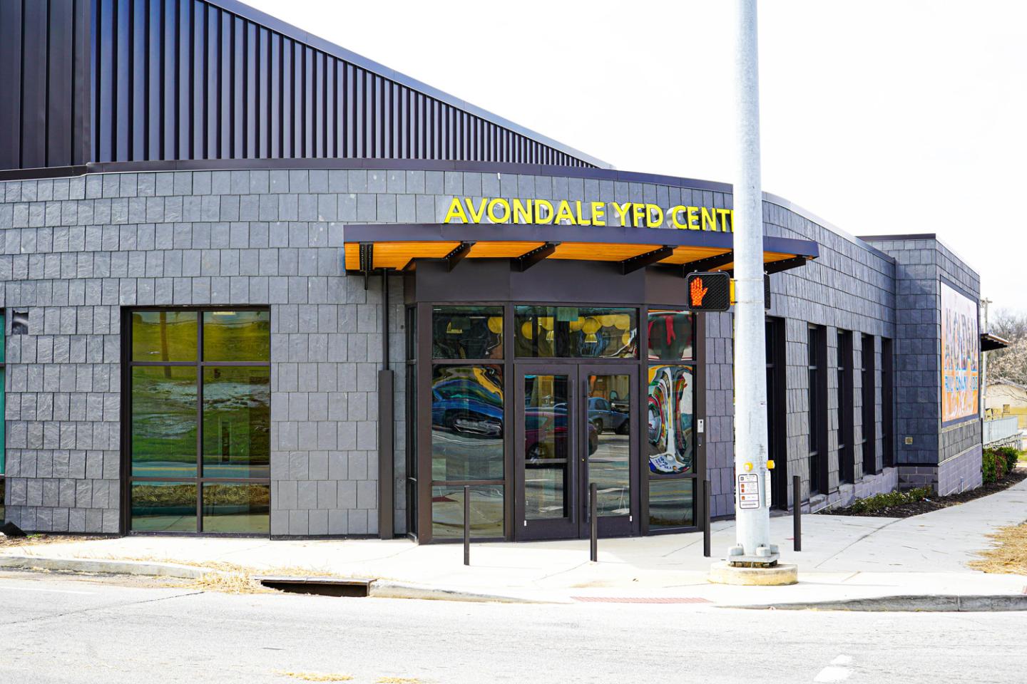 Facade of Avondale Community Center with gray brick walls, large glass doors, and a bright yellow sign under a wooden awning