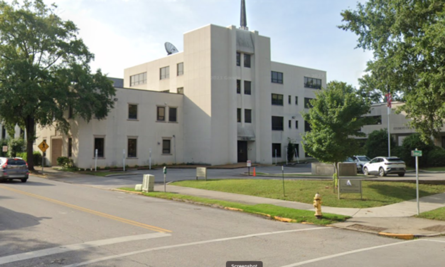 White multi-story building of First Centenary Community Center with a central tower and satellite dish, surrounded by trees and parked cars, on a sunny day