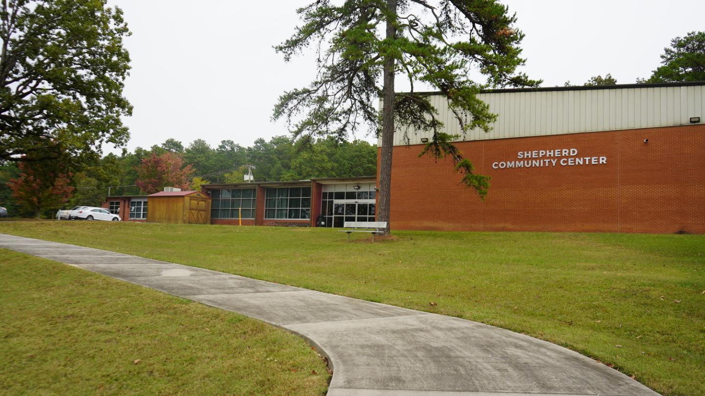 Exterior view of Shepherd Community Center with red brick walls and beige upper paneling, surrounded by green grass and trees, featuring a long concrete pathway leading up to the entrance.