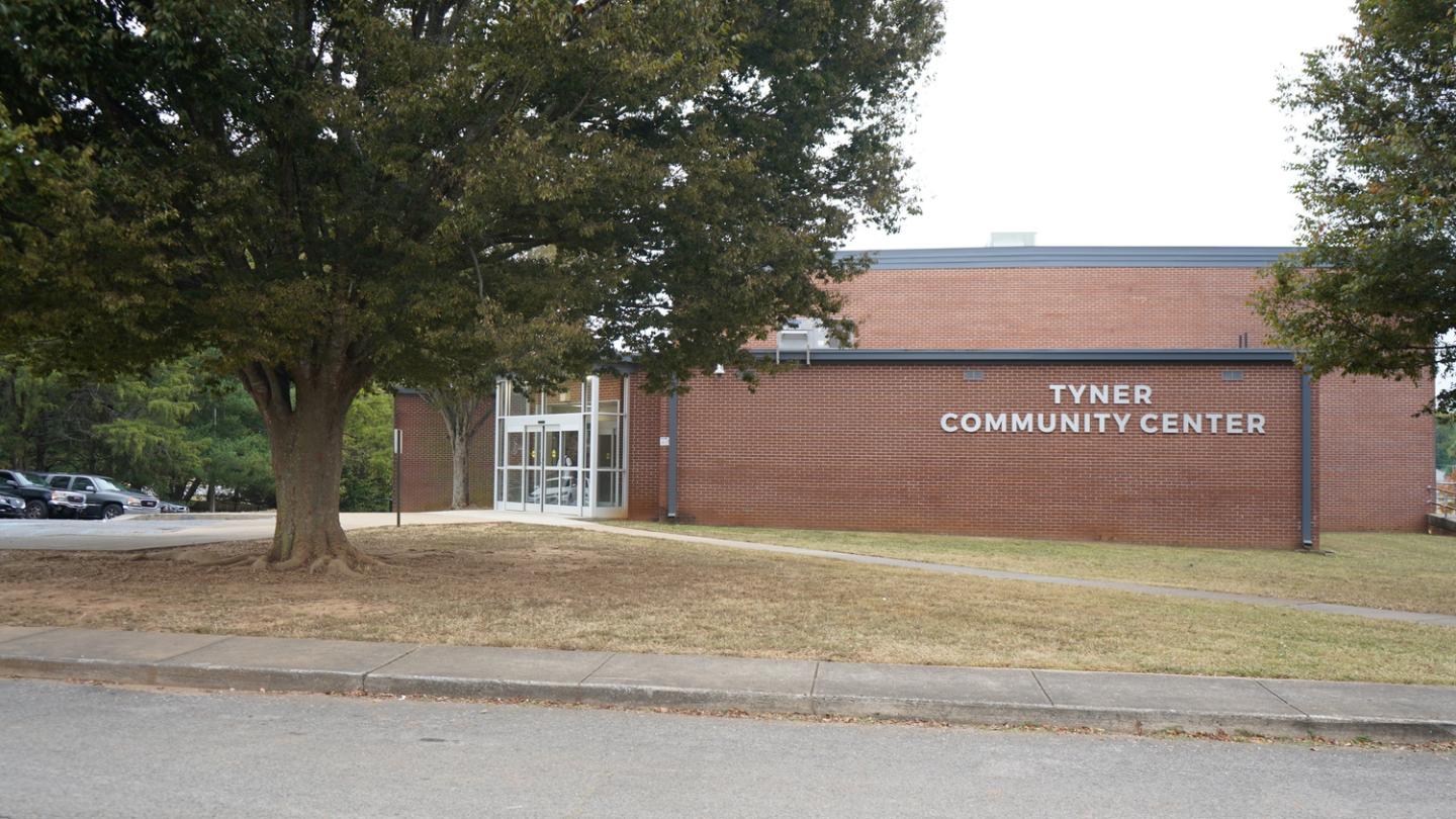 Red brick building of Tyner Community Center with large white letters, surrounded by green trees and a grassy area, with a clear glass entrance on the left side.