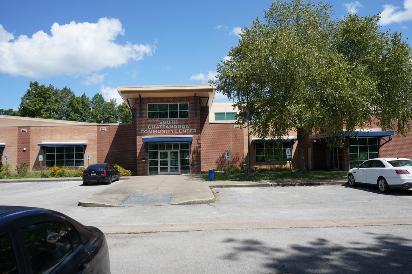 Exterior view of South Chattanooga Community Center on a sunny day, featuring a red brick building with a modern entrance, large green windows, and parked cars in the foreground. A large tree with green leaves is on the right side of the image