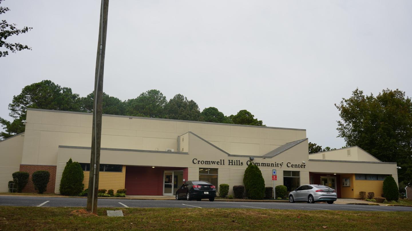 Exterior view of Cromwell Hills Community Center with beige and yellow walls, green bushes, and two parked cars in front of the building on a cloudy day