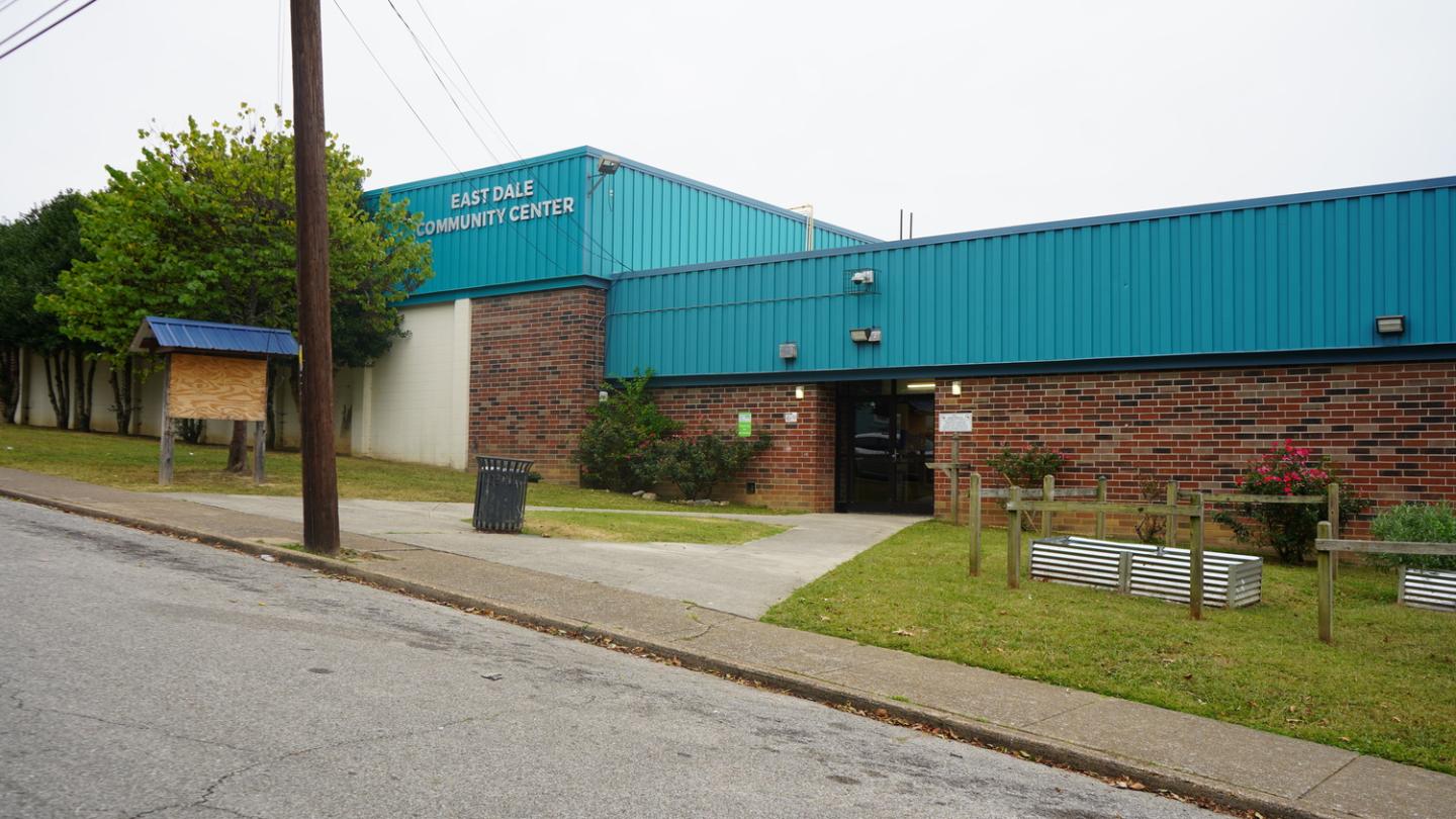 Front exterior view of Eastdale Community Center with blue and teal metal siding, brick walls, and a well-kept lawn featuring small plants and a wooden notice board