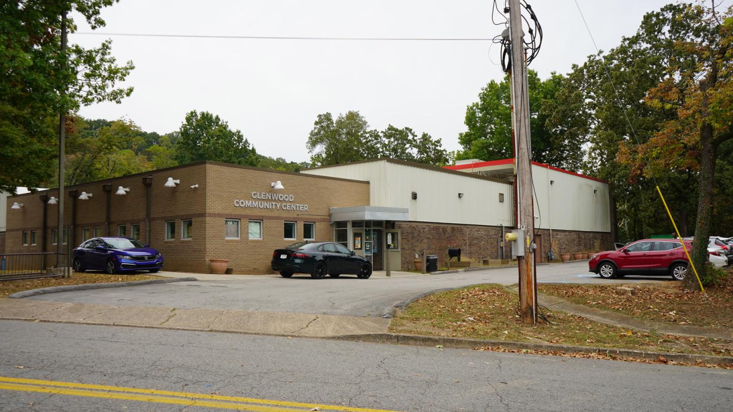 Glenwood Community Center, a light brown brick building with white accents, surrounded by green trees, with a red-roofed extension and multiple parked cars.