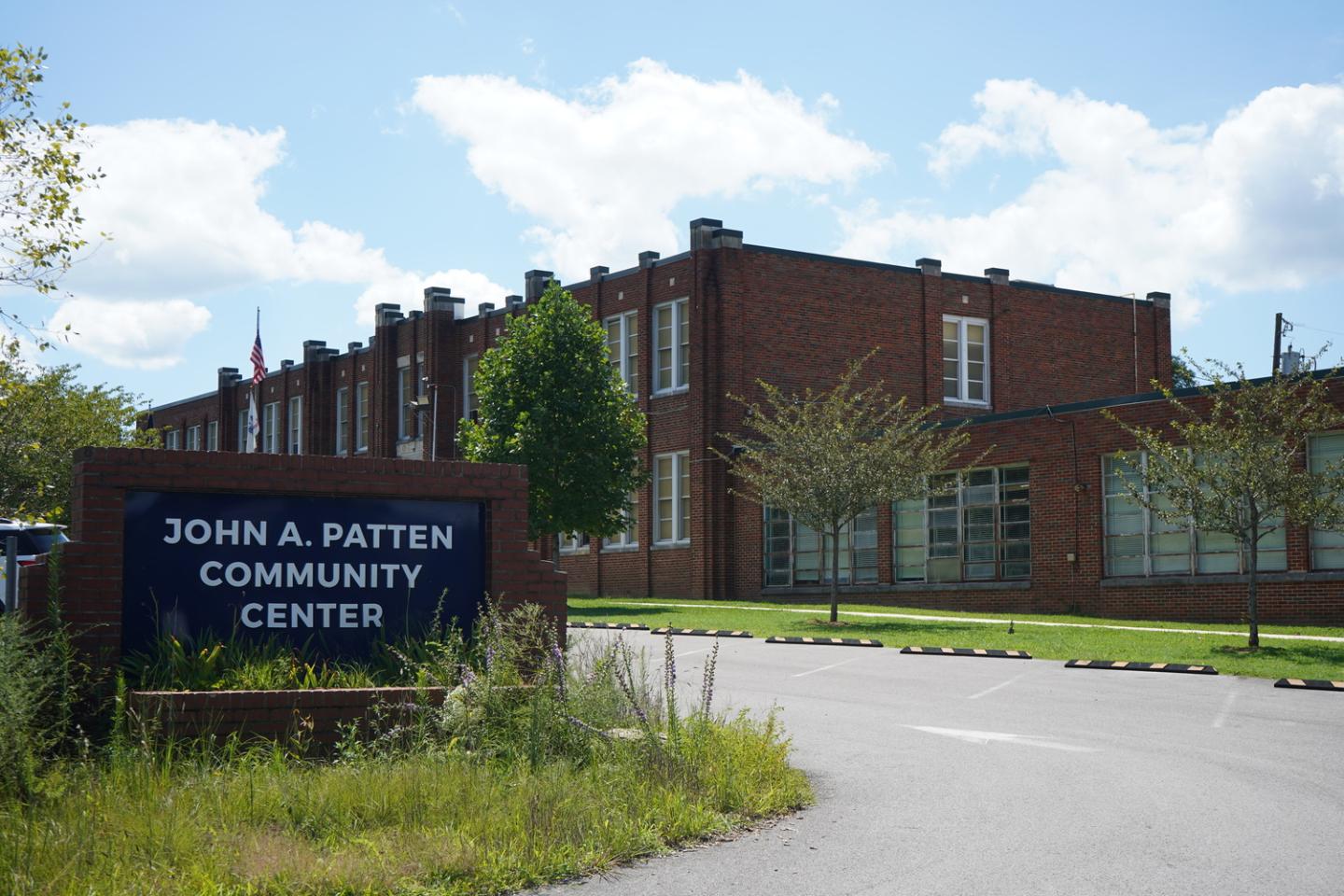 Brick building with white-trimmed windows under a blue sky, with a sign reading 'John A. Patten Community Center' in white letters on a dark blue background, surrounded by greenery