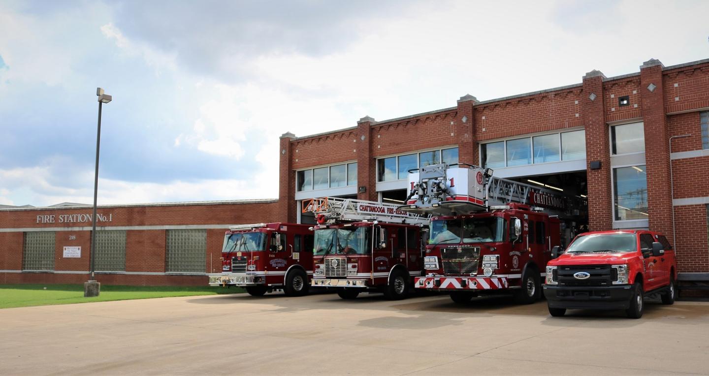 Three fire trucks and one red car parked in front of a brick building at fire station 1.