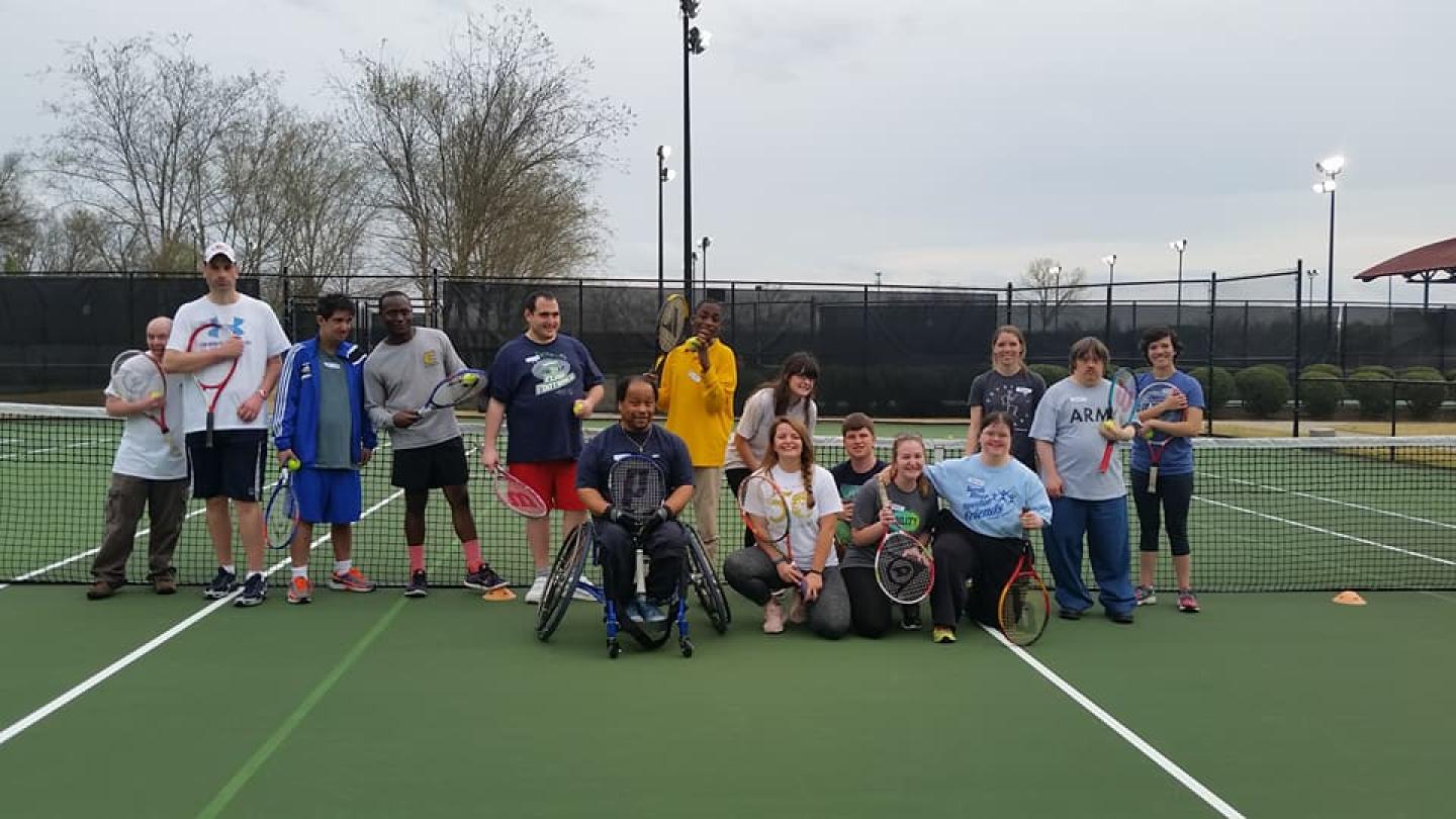 Group of people, including individuals in wheelchairs, holding tennis rackets on an outdoor tennis court.