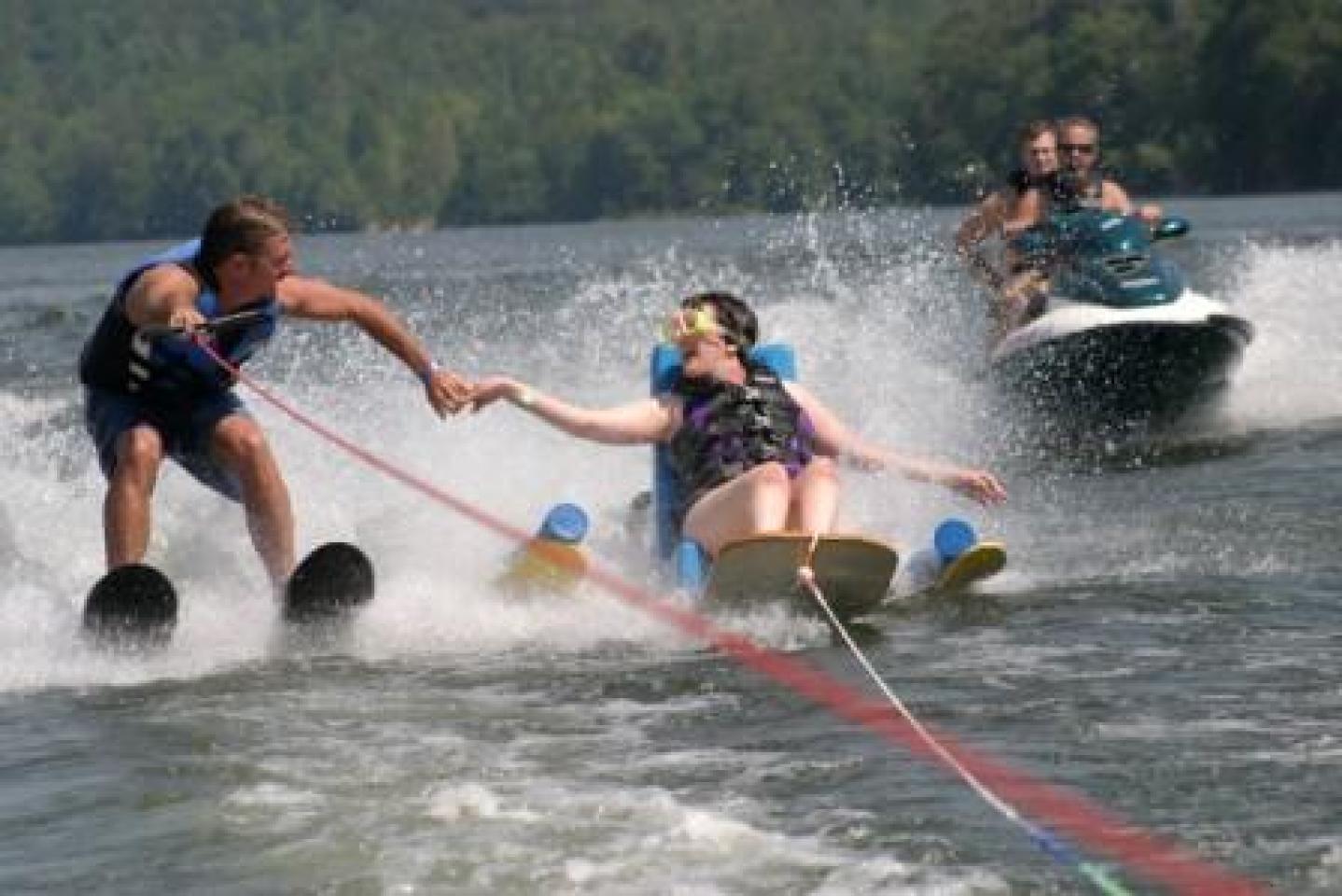 Man on water skis holding hands with a woman on adaptive water skis, assisted by a person on a jet ski in a lake setting