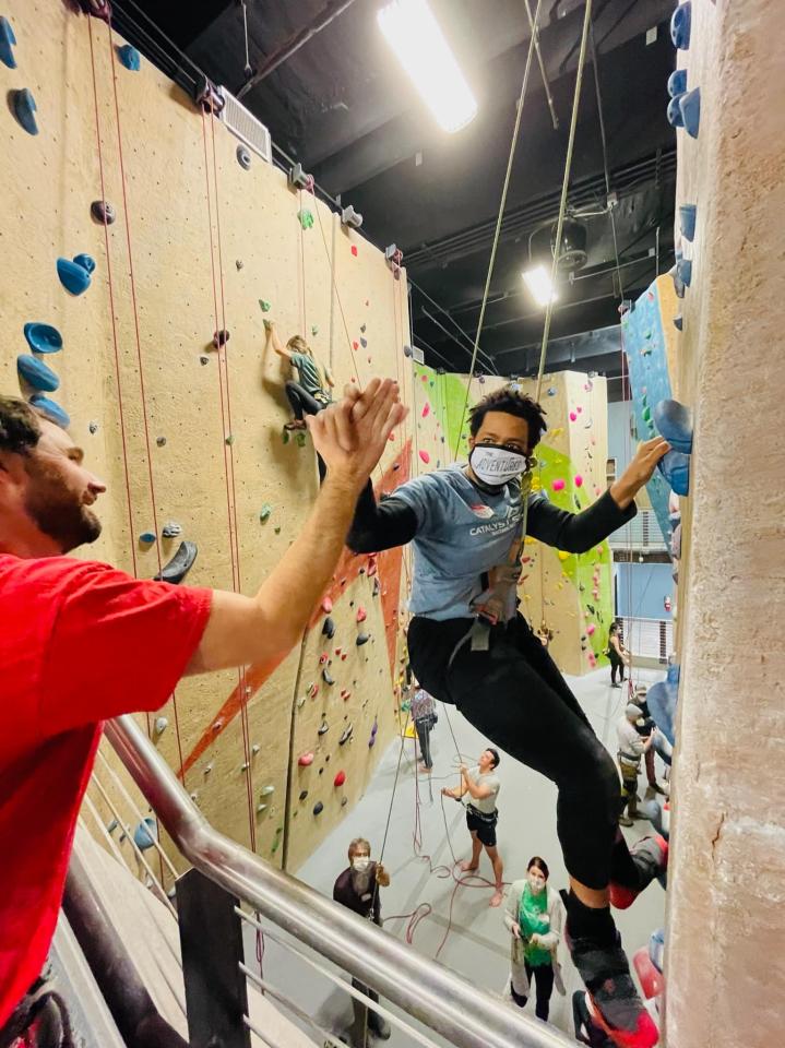 Person with a mask climbing an indoor rock wall, giving a high five to another climber in a red shirt