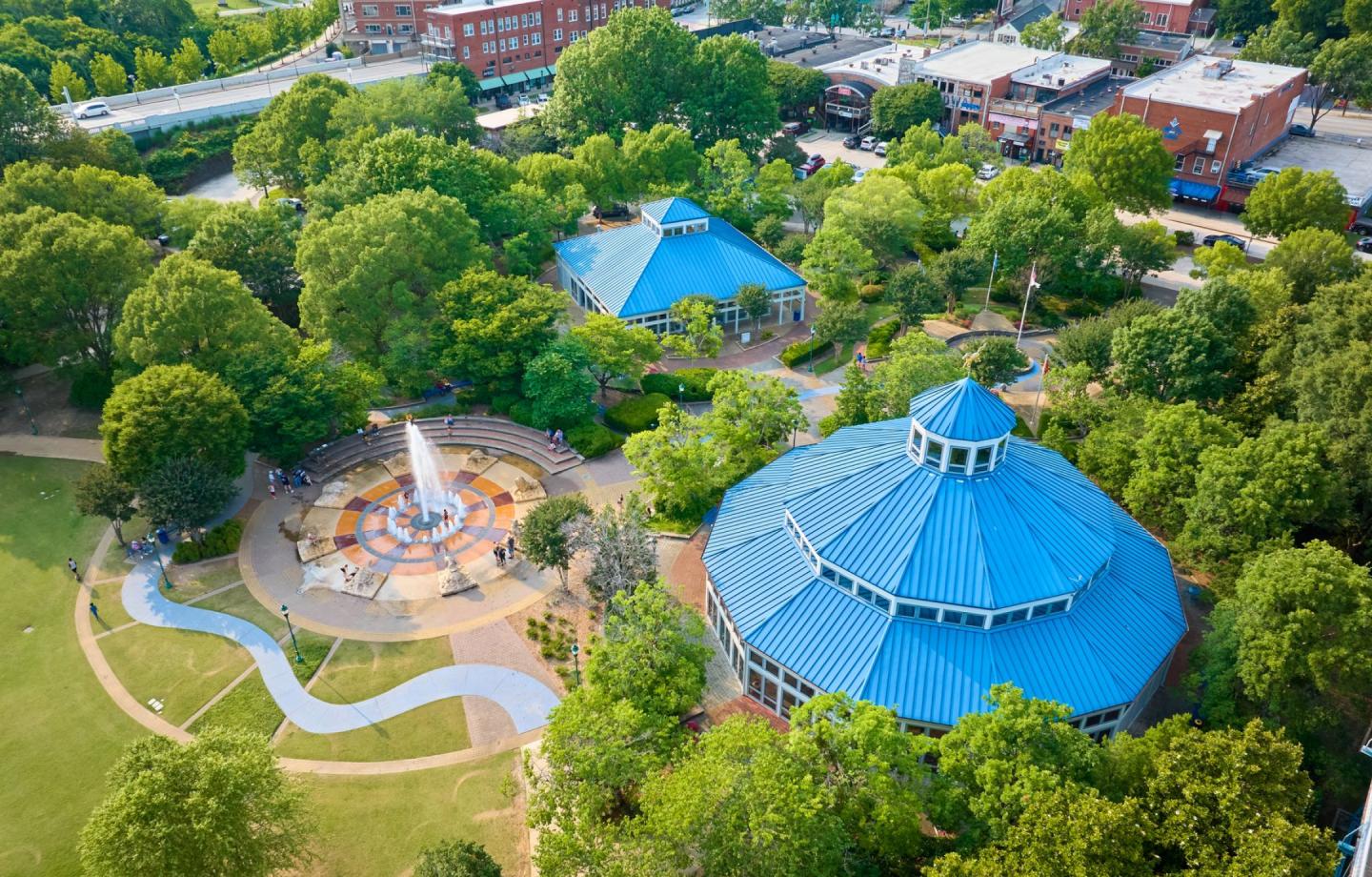 Aerial view of Coolidge Park with a carousel pavilion, fountain, and green spaces surrounded by trees and adjacent buildings.