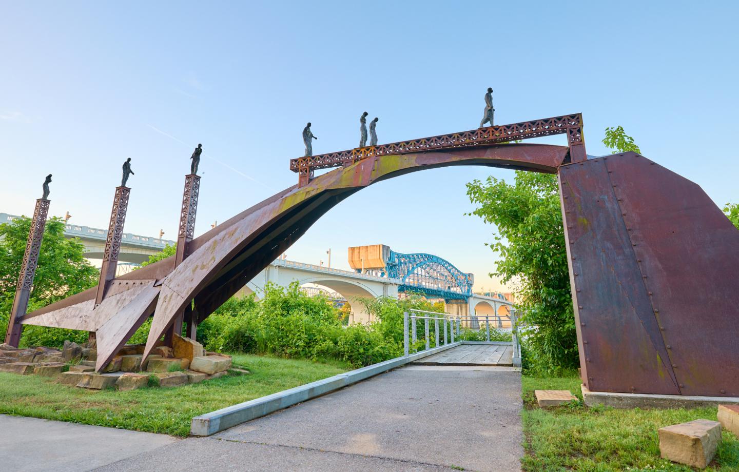 Sculptural bridge at Renaissance Park with human statues overlooking a pathway, greenery, and a distant blue bridge in Chattanooga, Tennessee.