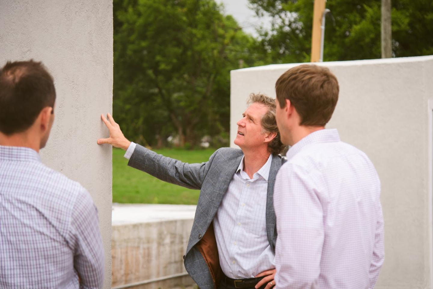 Three men standing in front of a building, wearing suits and smiling.