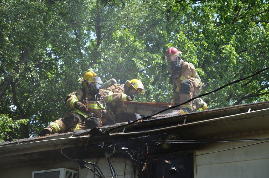 Firefighters on roof of house battling a fire, smoke billowing from the roof.