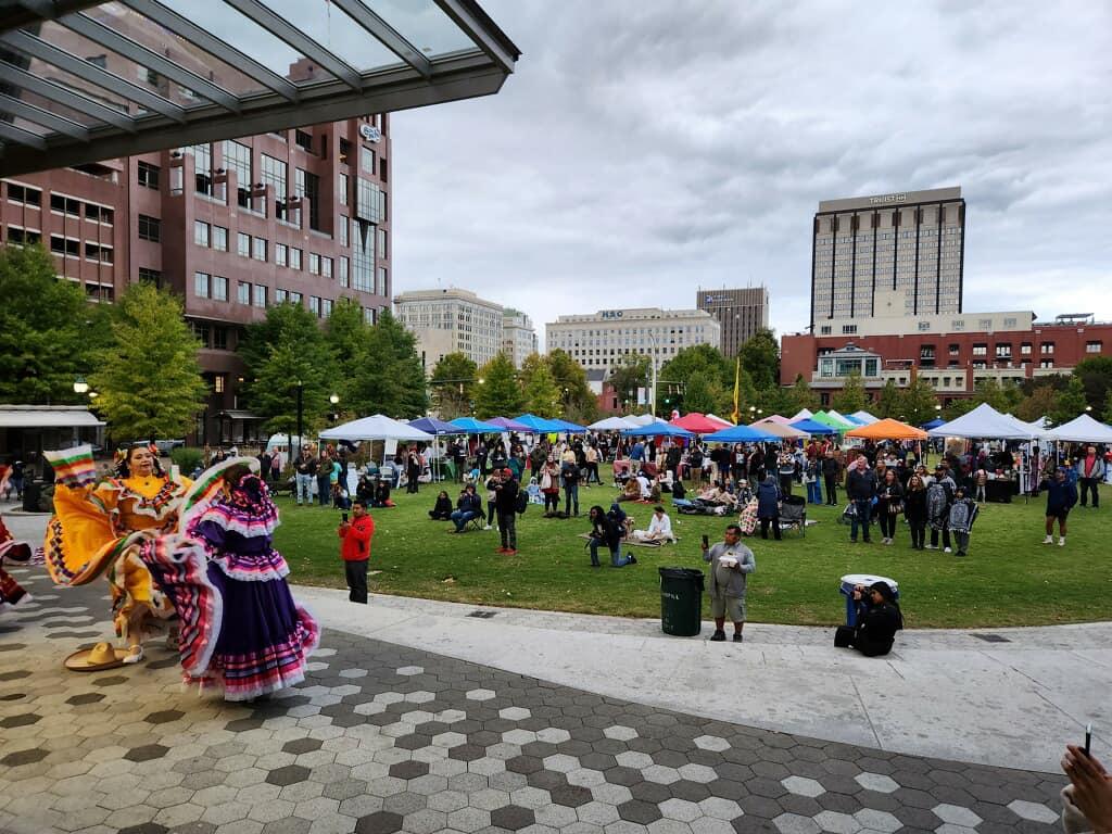 Outdoor festival in a city park with people enjoying various stalls and dancers in traditional attire