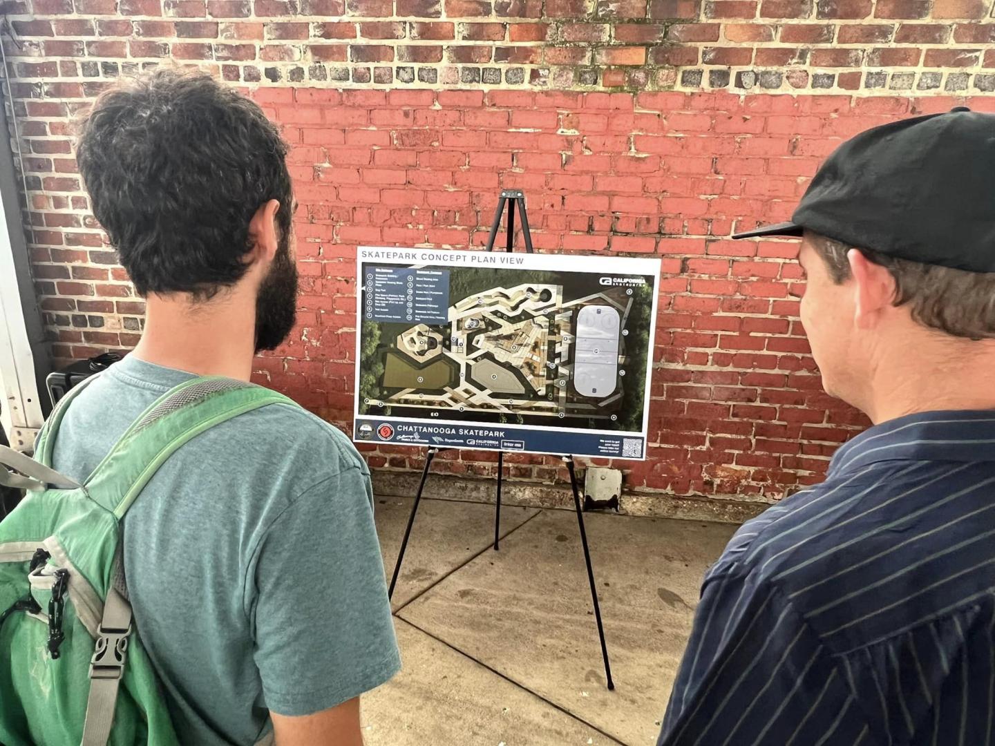 Two people are standing in front of an easel with a detailed map of a proposed skatepark pinned to it. They appear to be discussing the layout. The scene is set against a background of a red brick wall. One person is wearing a backpack.