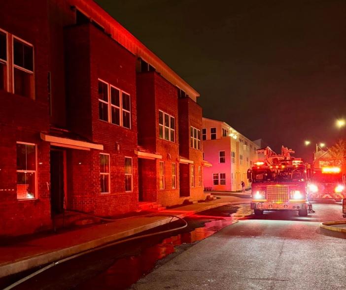 A fire truck with emergency lights on is parked outside a multi-story residential building at night. The building's exterior is illuminated by the red lights. The scene appears to be calm with no visible flames or smoke.