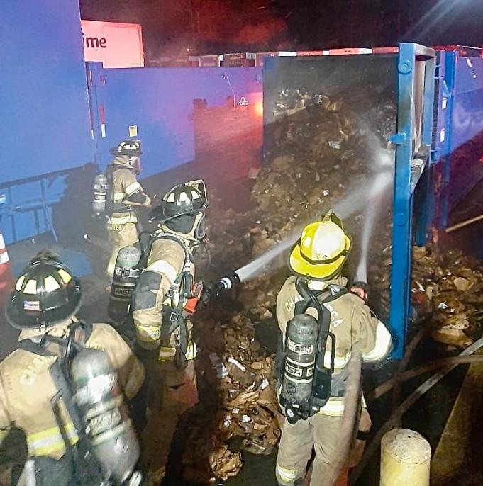 Four firefighters in full gear and oxygen tanks are using hoses to extinguish a fire in a large blue dumpster filled with cardboard boxes and debris. It is nighttime, and the area around them is dimly lit by streetlights and the glow of the fire.