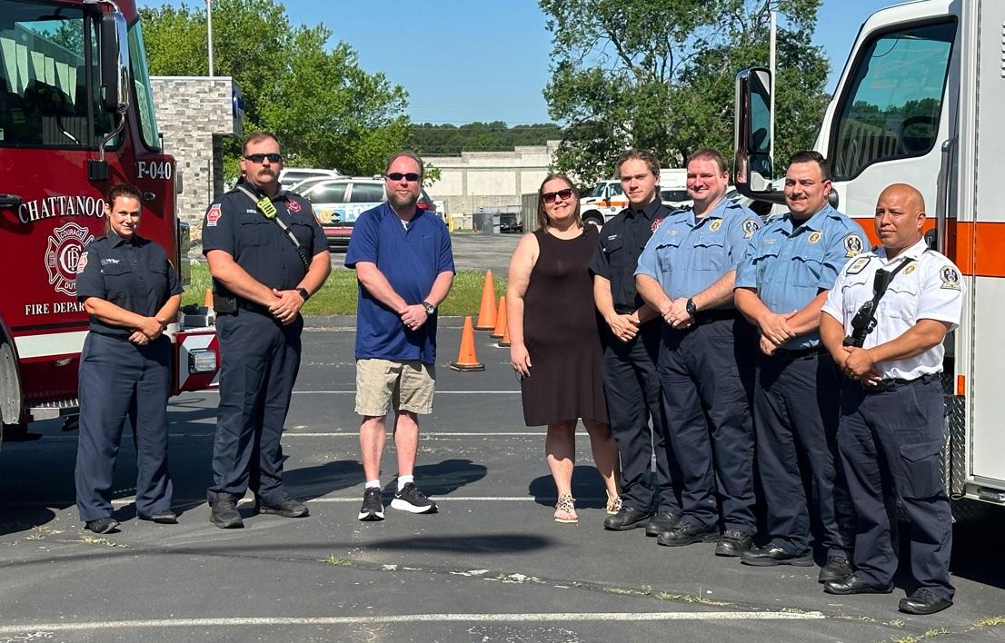 A group photo shows seven emergency service personnel standing next to a fire truck and an ambulance. Four men and three women are dressed in various uniforms, including firefighter and paramedic attire. Two people in civilian clothes stand in the middle of the group.