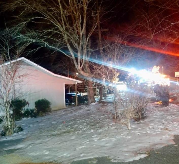 A small house is pictured at night with bright lights illuminating the scene. The ground is partially covered with snow, and leafless trees stand in the yard. The surrounding area suggests a cold, possibly winter evening.