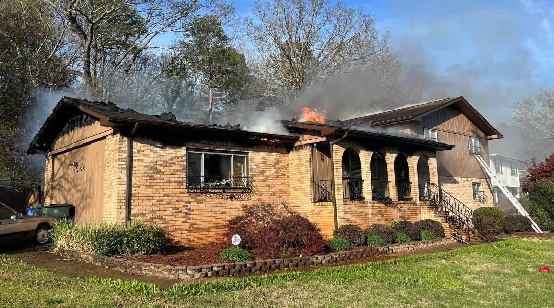 A single-story brick house with an attached multi-story structure is on fire, with smoke and flames visible on the roof. Trees without leaves are in the background, and the front yard features various shrubs and plants. A staircase leads to the multi-story section.