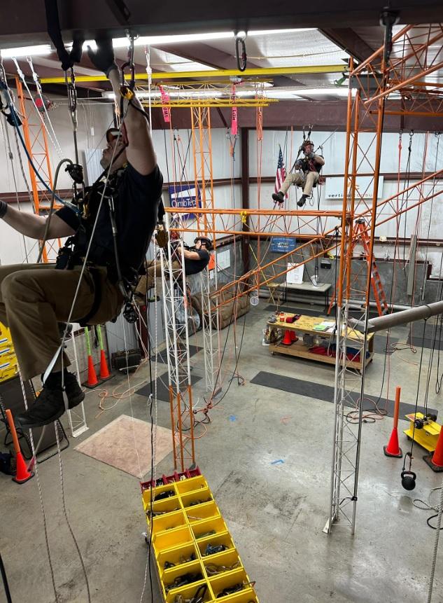 A group of people participate in a rope access training session inside an industrial facility. They are suspended from orange and white metal structures, surrounded by safety cones, ropes, and equipment storage bins. American flags are visible in the background.