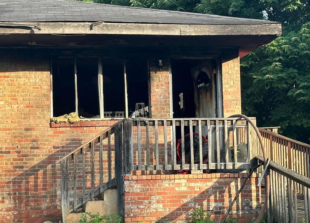 Image of a brick house with significant fire damage. The windows are broken, and the surrounding walls are charred. The wooden front steps and railing remain intact but show signs of wear. The front door is partially open, revealing more damage inside. Trees are in the background.