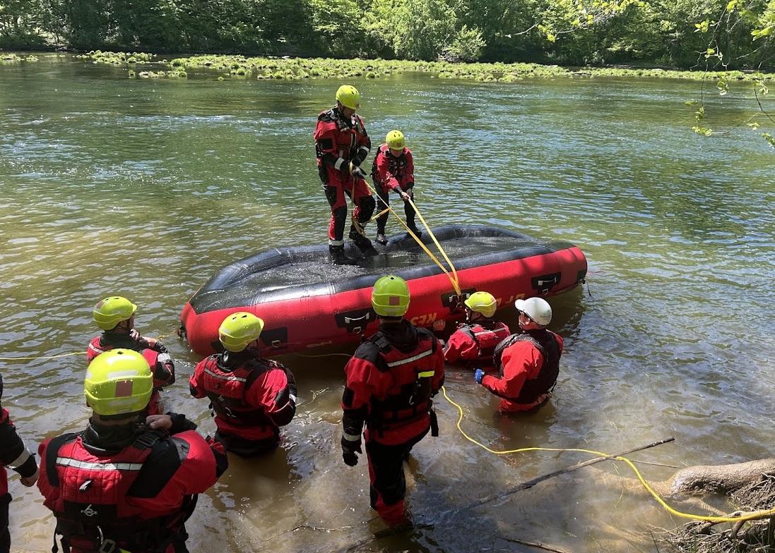 Rescue workers in red and black outfits and yellow helmets conduct a training exercise on water rescue. Some are on an inflated raft while others are in water or on the shore, holding onto ropes. The background includes trees and a partly sunlit river.