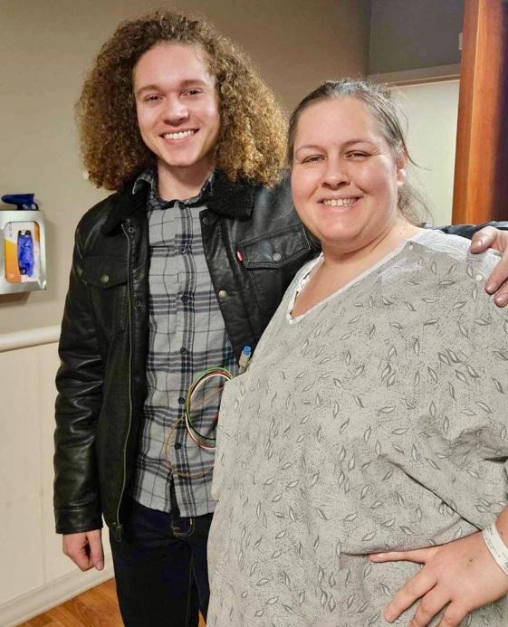 A young man with curly hair, wearing a leather jacket, and a woman with long hair, smiling and posing together in an indoor setting. The woman is wearing a grey patterned top and has her arm around the man's shoulder.