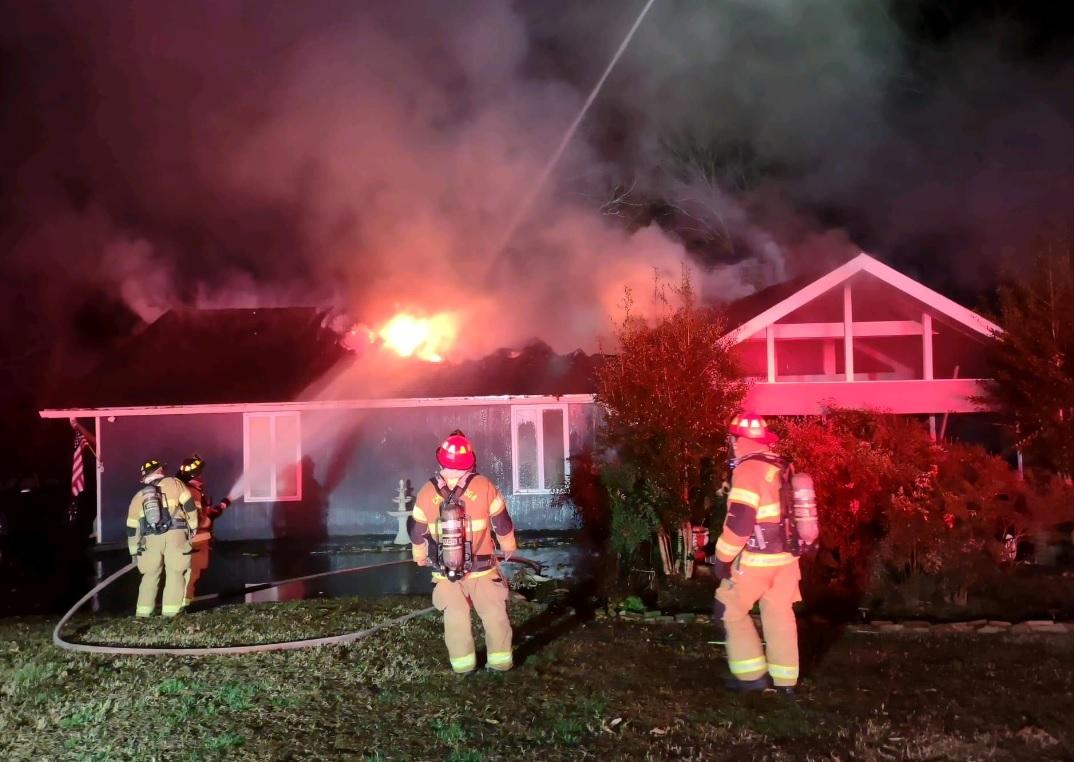 Firefighters in protective gear battle a house fire at night. Flames and smoke are seen coming from the roof as one firefighter uses a hose. The scene is illuminated by fire engine lights. The house appears to be a single-story residence with significant fire damage.