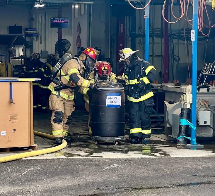 Three firefighters in full gear and breathing apparatus inspect a black barrel labeled "Copper Dust" inside an industrial workshop. The scene includes various equipment and machinery, a large box on the left, and coiled hoses hanging from the ceiling.