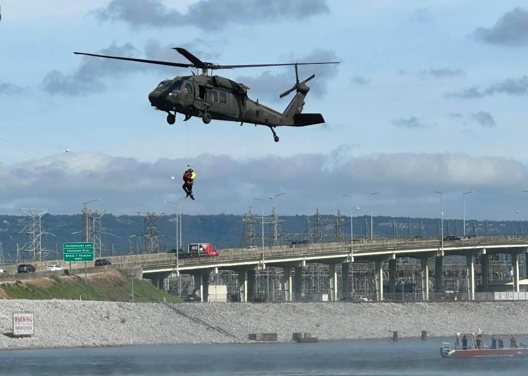 A helicopter hovers above a body of water with a person suspended underneath, likely during a rescue operation. In the background, there's an industrial area with power lines and a bridge spanning across the water. A boat is seen near the water's edge.