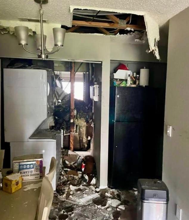 A room with severe structural damage showing an exposed ceiling with broken drywall. Debris is scattered across the room. A washing machine and dryer unit is visible amidst the damage, and a damaged electrical panel is on the right wall. A chandelier hangs from the ceiling.