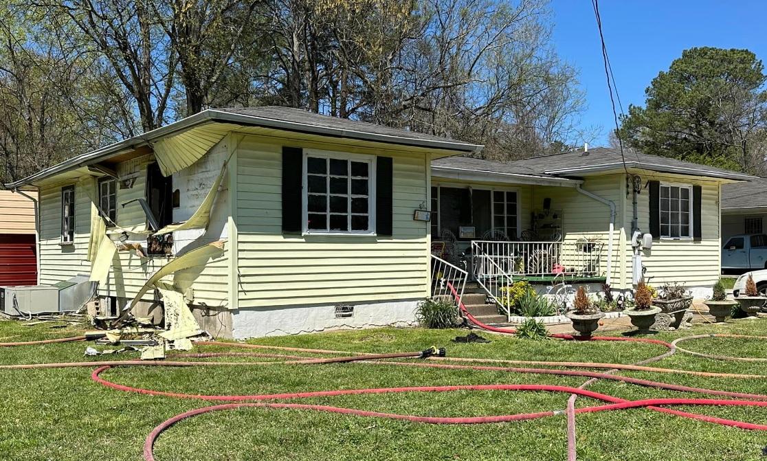 A small, yellow house with visible fire damage on the exterior. The siding is melted, and charred marks are present around the windows. Hoses are sprawled across the lawn, indicative of recent firefighting efforts. The surrounding trees and blue sky are visible.