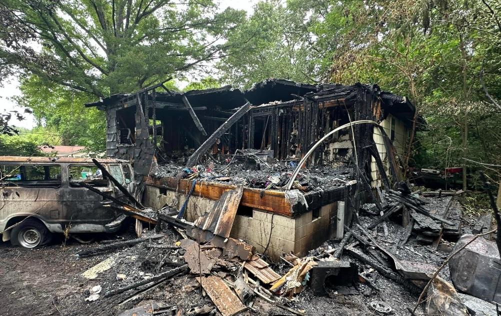 A fire-ravaged house almost entirely destroyed, with charred debris and burnt wood scattered around. The blackened remains of the structure are surrounded by tall trees, and a severely damaged vehicle is parked on the left. The scene shows extensive damage and destruction.