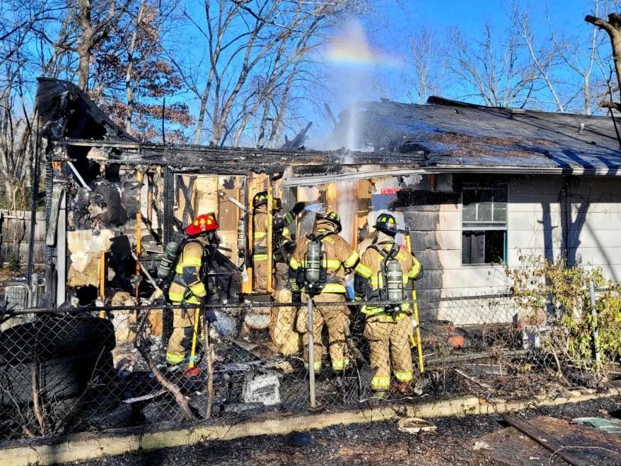 Firefighters wearing gear and helmets work together to extinguish the remains of a house fire. The house's roof and walls are badly burned, and smoke is rising from the structure. A hose sprays water on the charred roof beams, and a faint rainbow can be seen.