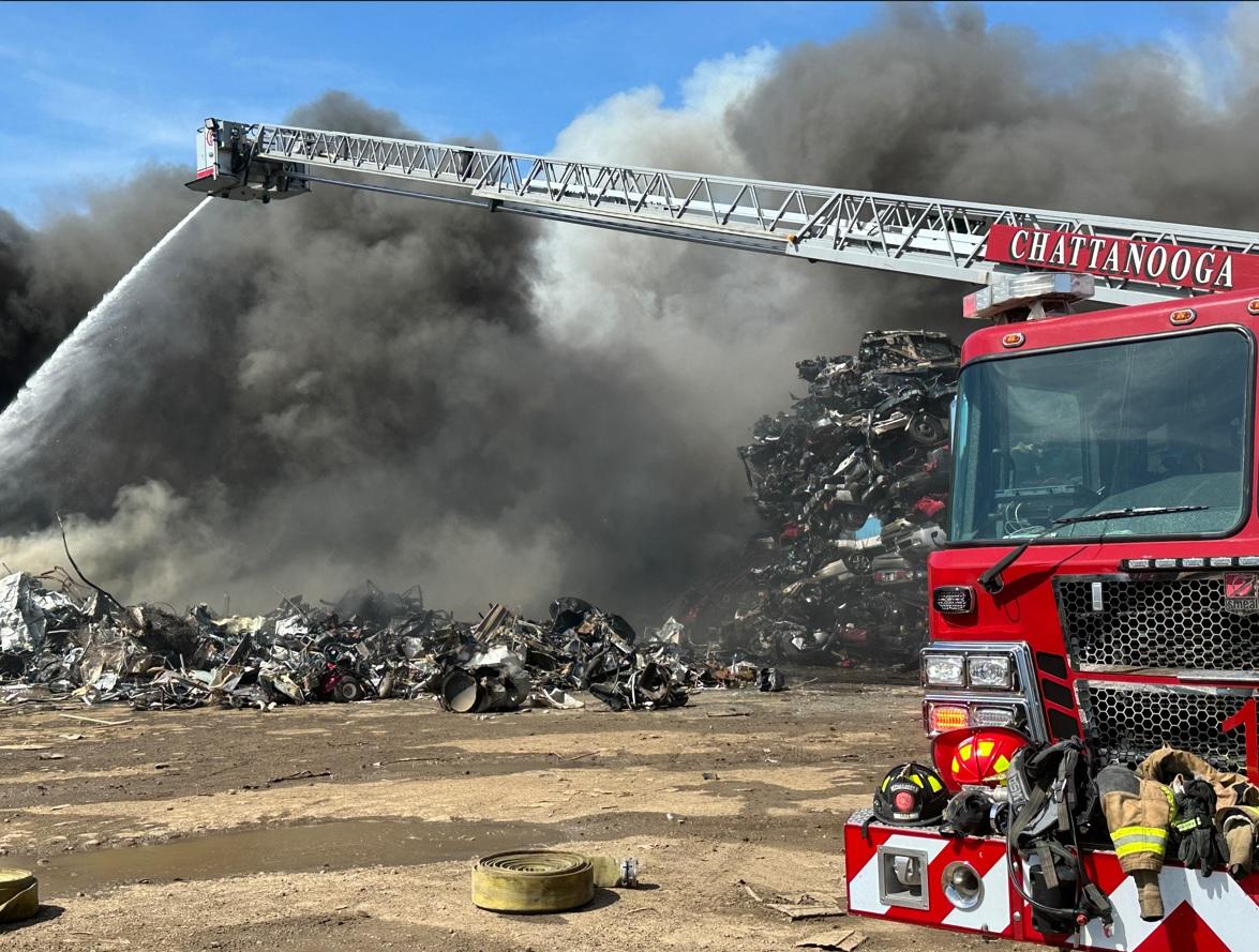 A large fire truck with "Chattanooga" on its side extends its ladder spraying water onto a large pile of metal scrap engulfed in thick black smoke. Debris is scattered on the ground, and the sky is clear blue in the background.