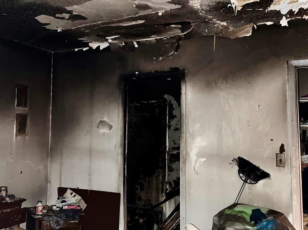 A room with significant fire damage. The ceiling has large holes and is blackened from soot. The walls are charred, especially around a doorway leading to another damaged area. Debris and clutter are scattered on the floor and a unburnt broom leans in a corner.