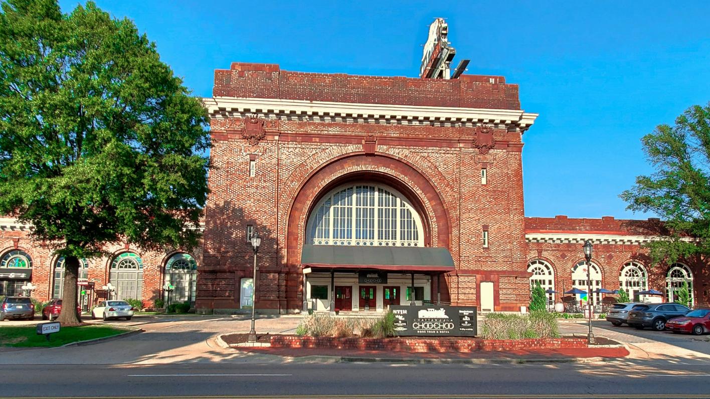 Front view of the historic Chattanooga Choo Choo Terminal Station with its distinctive red brick architecture, large arched window, and surrounding greenery on a clear, sunny day.